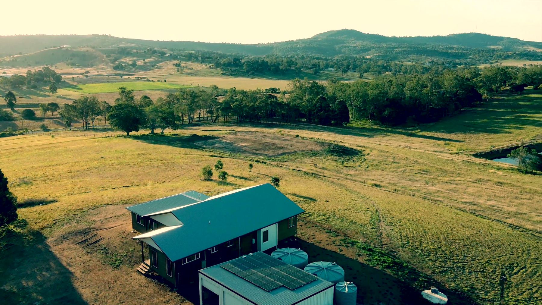 Aerial view of an off-grid home in South East Queensland, with solar panels on the shed roof and water tanks, and hills behind